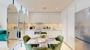 A modern kitchen with white cabinets, a dining table set with plates, glasses, and cutlery, and a green sofa in the adjacent living area. - Luxe 1 Bed l Private Beach Access & City Views (Emaar Beachfront)
