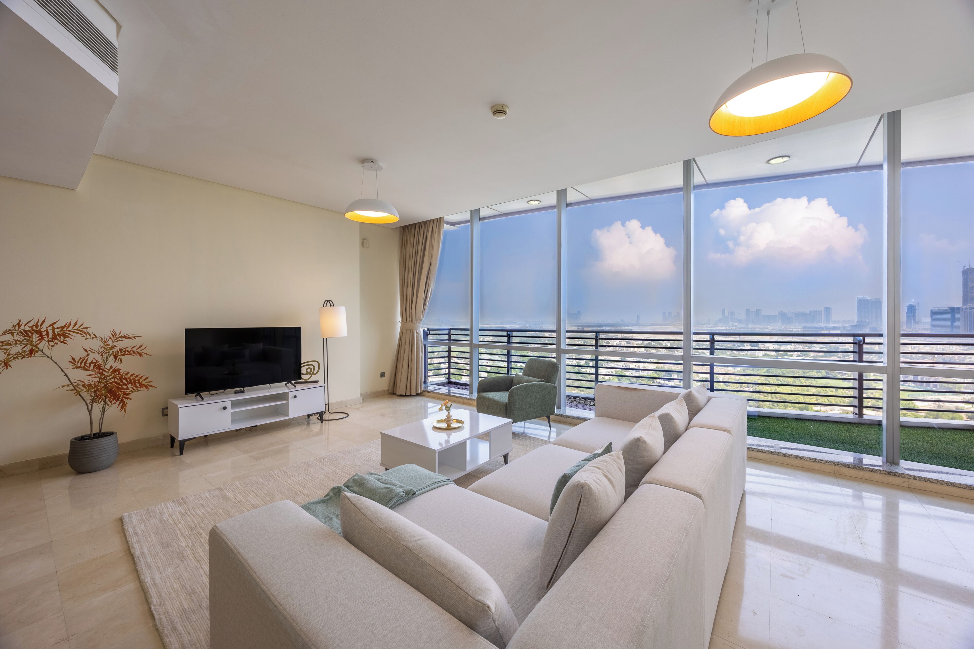 A modern living room with a large sofa, a flat-screen TV, a potted plant, and a view of a city skyline through large windows.
