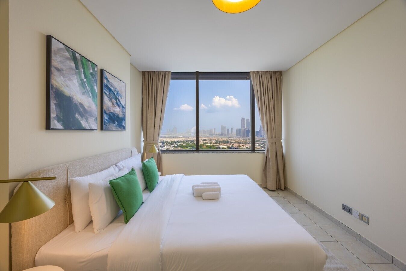 A hotel room with a large bed, two pillows, a lamp, and a view of a cityscape through a window.
