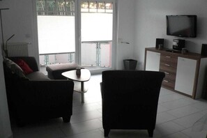Un salon avec un canapé, un fauteuil et une petite table. On y trouve un téléviseur posé sur un meuble et une commode avec des tiroirs.