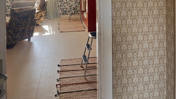 A room with patterned wallpaper, a radiator, a window with curtains, a chair, a ladder, and a red door.