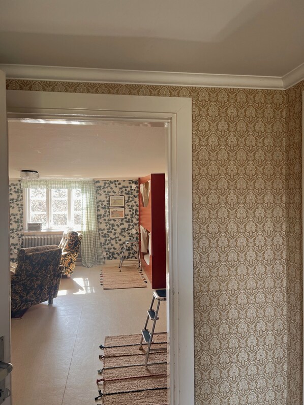 A room with patterned wallpaper, a radiator, a window with curtains, a chair, a ladder, and a red door.