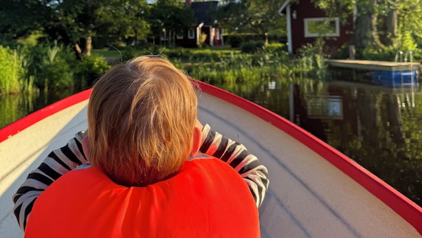 Henkilö veneessä, päällään punainen pelastusliivi, veden ja puiden ympäröimänä, taustalla näkyy talo.