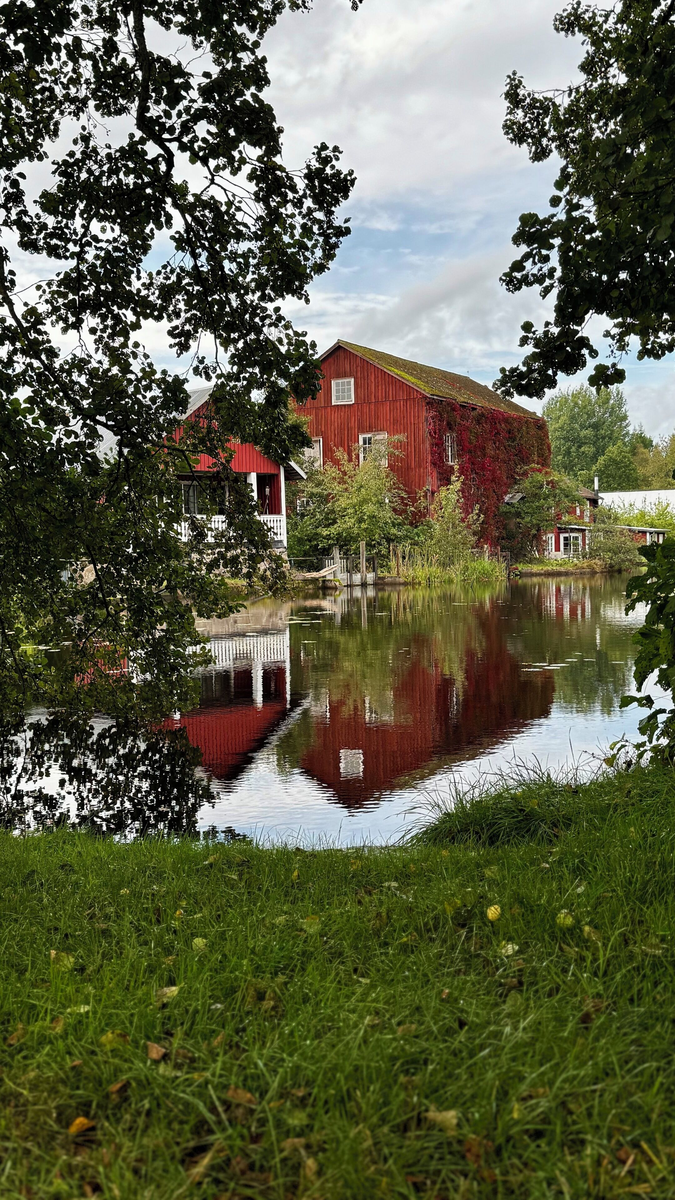 En rød lade med et spejlbillede i stille vand, omgivet af grøn vegetation.