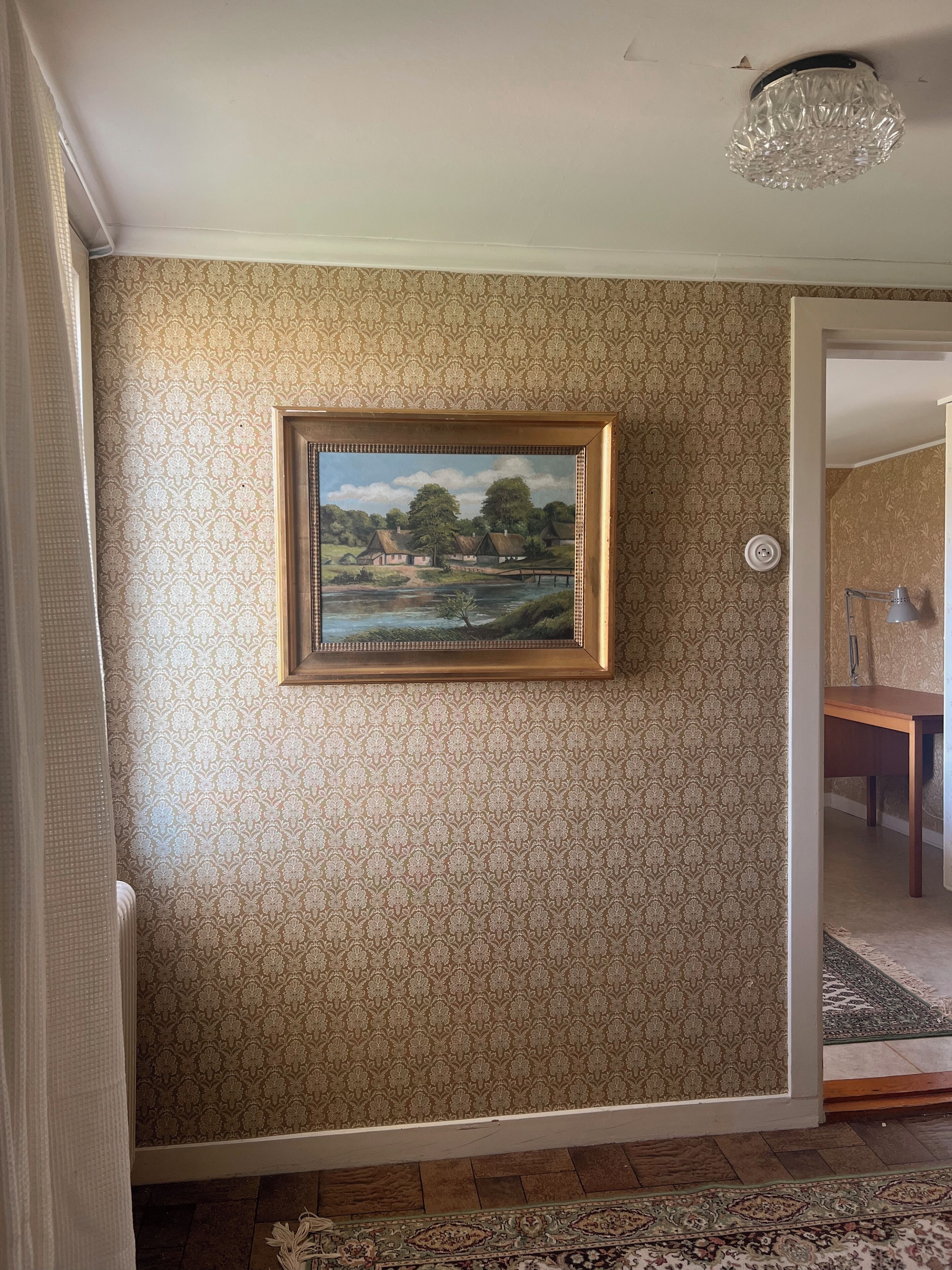 Une pièce avec un papier peint à motifs, un tableau représentant un paysage encadré, un lustre et une table en bois.