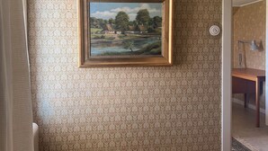 A room with patterned wallpaper, a framed landscape painting, a chandelier, and a wooden table.