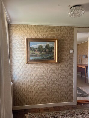 A room with patterned wallpaper, a framed landscape painting, a chandelier, and a wooden table.