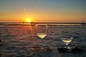 Zwei Gläser Weißwein an einem Sandstrand, im Hintergrund geht die Sonne unter.