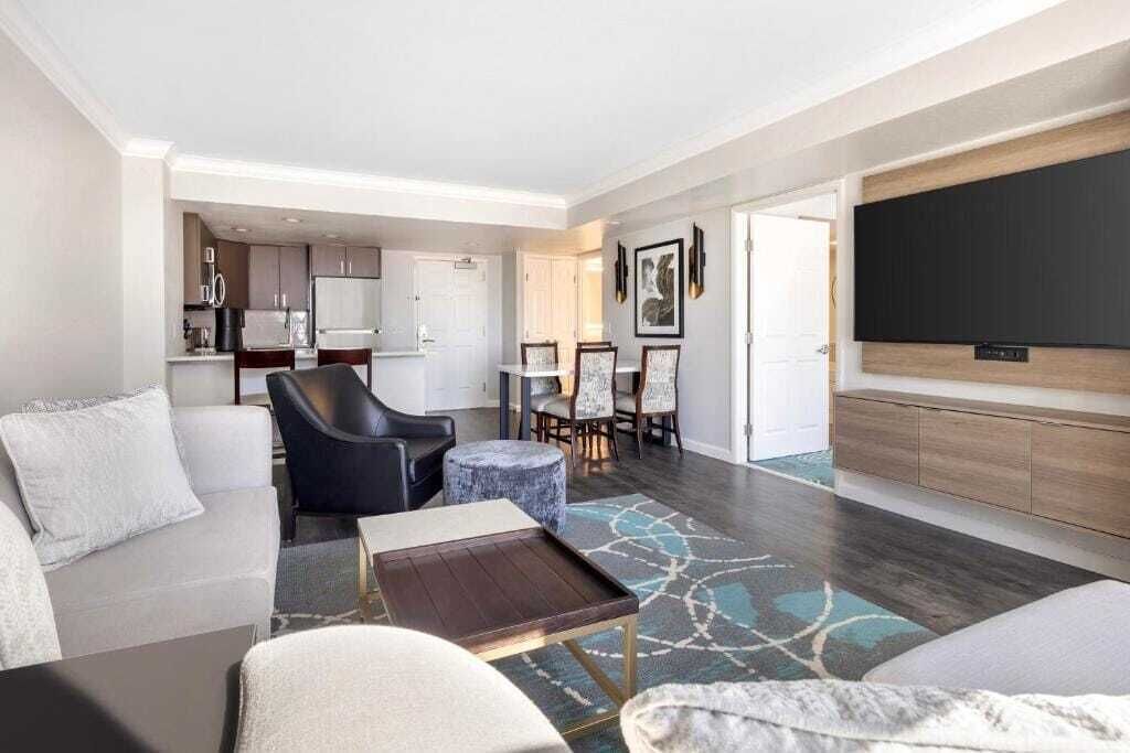 A modern living room with a sofa, a coffee table, and a television. There is a dining area with a table and chairs. The room has a neutral color palette and a patterned rug.