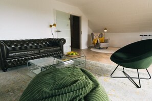 Ein Wohnzimmer mit einer Ledersofa, einem grünen Sessel, einem Glastisch und einer Tür zu einem weiteren Raum.