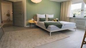 Quarto com cama, mesas de cabeceira, luminária pendente e vista para o exterior pela janela.