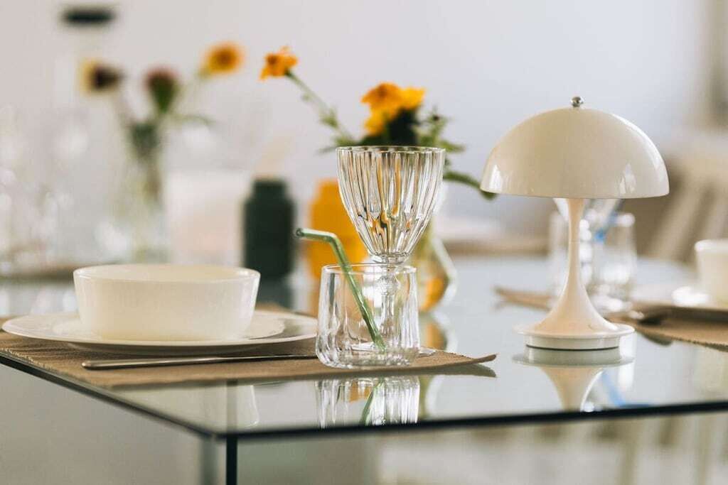 Una mesa de comedor con una lámpara de mesa blanca, un florero de vidrio transparente con un tallo verde y un cuenco blanco sobre un mantel individual.