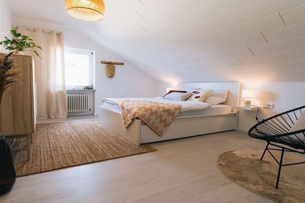 A modern bedroom with a sloped ceiling, a bed with a patterned throw, a bedside lamp, a chair, and a plant.