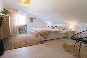 Ein modernes Schlafzimmer mit schrägem Dach, einem Bett mit gemustertem Überwurf, einer Nachttischlampe, einem Sessel und einer Pflanze.