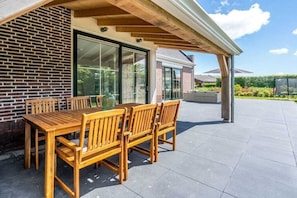 Een overdekt terras met een houten tafel en stoelen, een bakstenen muur en een uitzicht op een huis op de achtergrond.