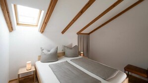 A bedroom with a sloped ceiling, a skylight, a bed with pillows, a bedside table with a lamp, and a bench. - Holiday apartment with 1 bedroom (Lindau (Bodensee))
