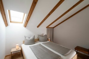 Ein Schlafzimmer mit schrägen Wänden, einem Oberlicht, einem Bett mit Kissen, einem Nachttisch mit Lampe und einer Bank.