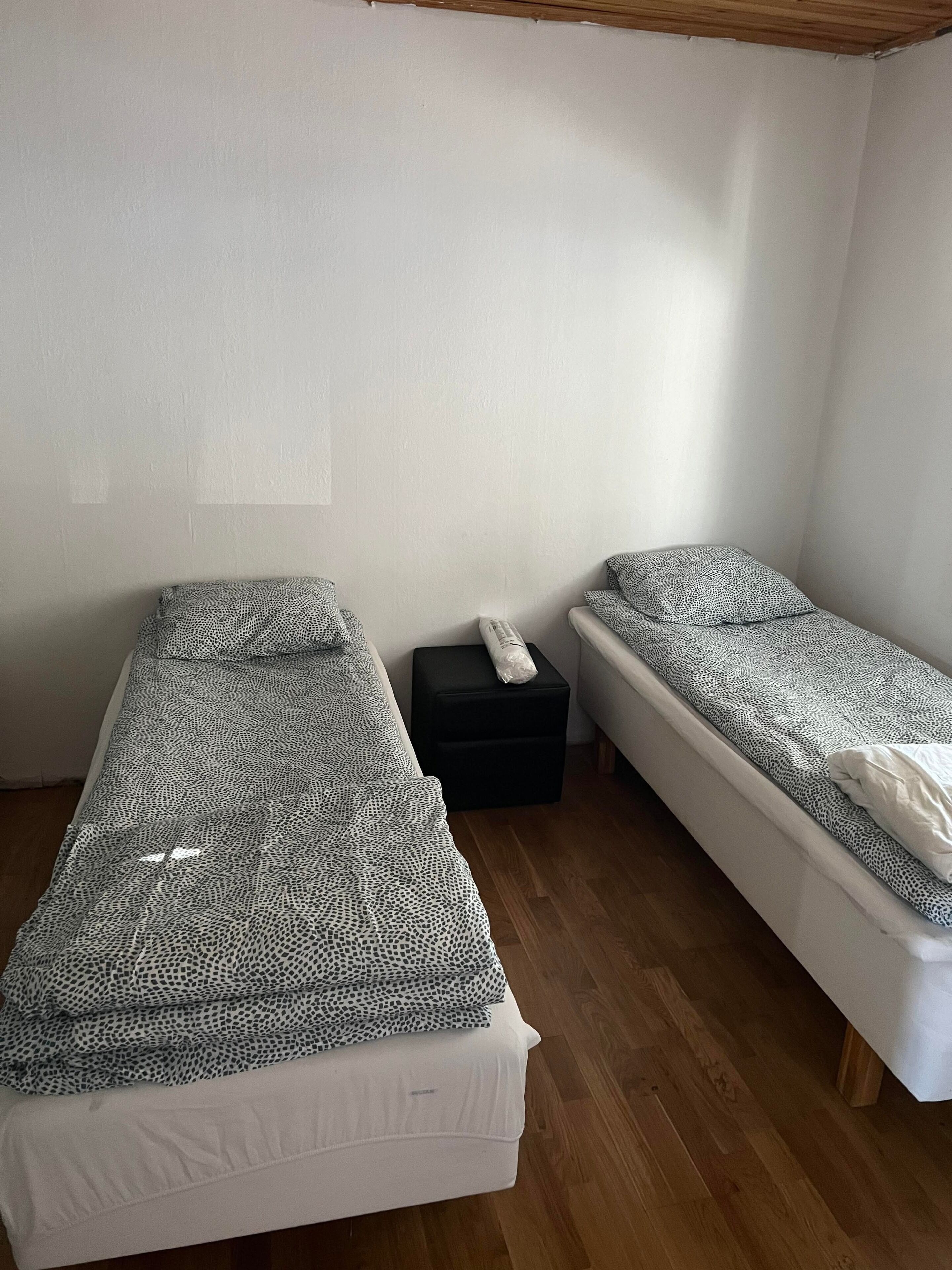 Una habitación con dos camas individuales, un pequeño taburete negro y piso de madera.
