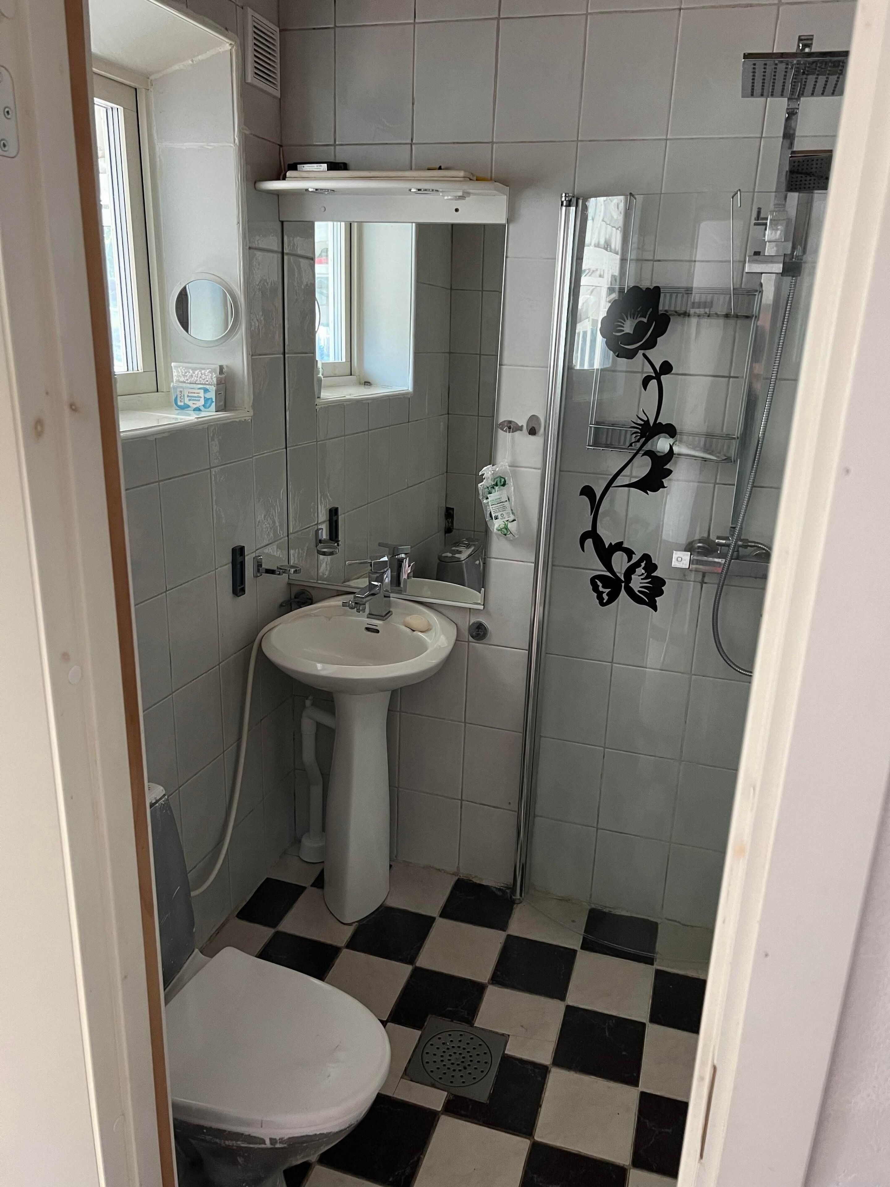 Une salle de bain avec un lavabo, des toilettes et une douche, le tout de couleur blanche. Un miroir se trouve au-dessus du lavabo et une fenêtre au-dessus des toilettes.