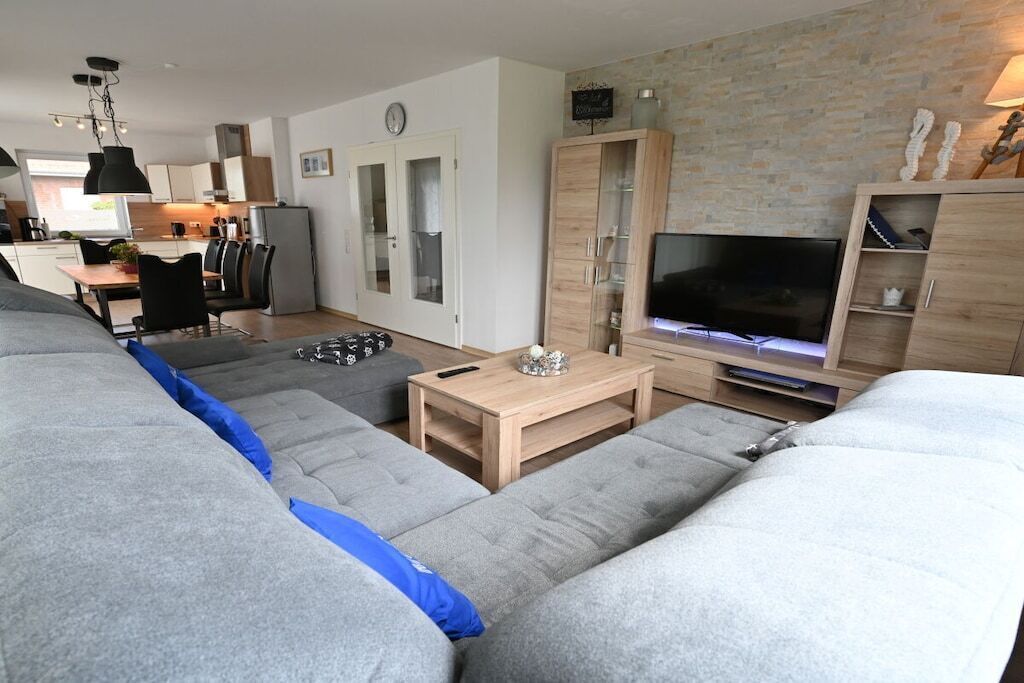 A modern living room with a sofa, a coffee table, and a TV. There is a kitchen in the background with cabinets and a refrigerator.