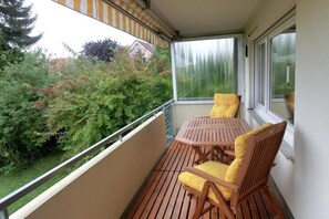 Een balkon met een houten tafel en stoelen, uitzicht op groen, en een raam.