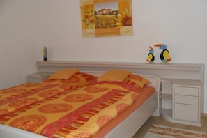 A bed with a patterned bedspread, a nightstand with drawers, a wall-mounted shelf with a painting, and two decorative bird figurines. - Holiday apartment with 1 bedroom (Meersburg)