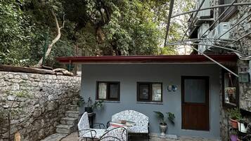 Un patio con pavimento in pietra, due poltrone decorate e un tavolo, un piccolo edificio con una porta e finestre, e un muro in pietra.