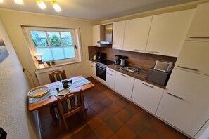 Eine kompakte Küche mit weißen Schränken, einem Esstisch mit Stühlen, einem Fenster mit Jalousien und einem eingebauten Backofen.
