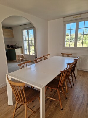 A dining area with a white table and wooden chairs, a kitchen in the background, and a window with a view of greenery. - A house at the foot of the beaches and the GR34 trail, in the heart of the Côte des Légendes. (Kerlouan)