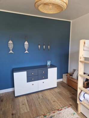 A room with a blue wall, a wooden floor, a white and dark blue cabinet with drawers, and a hanging light fixture. - A house at the foot of the beaches and the GR34 trail, in the heart of the Côte des Légendes. (Kerlouan)