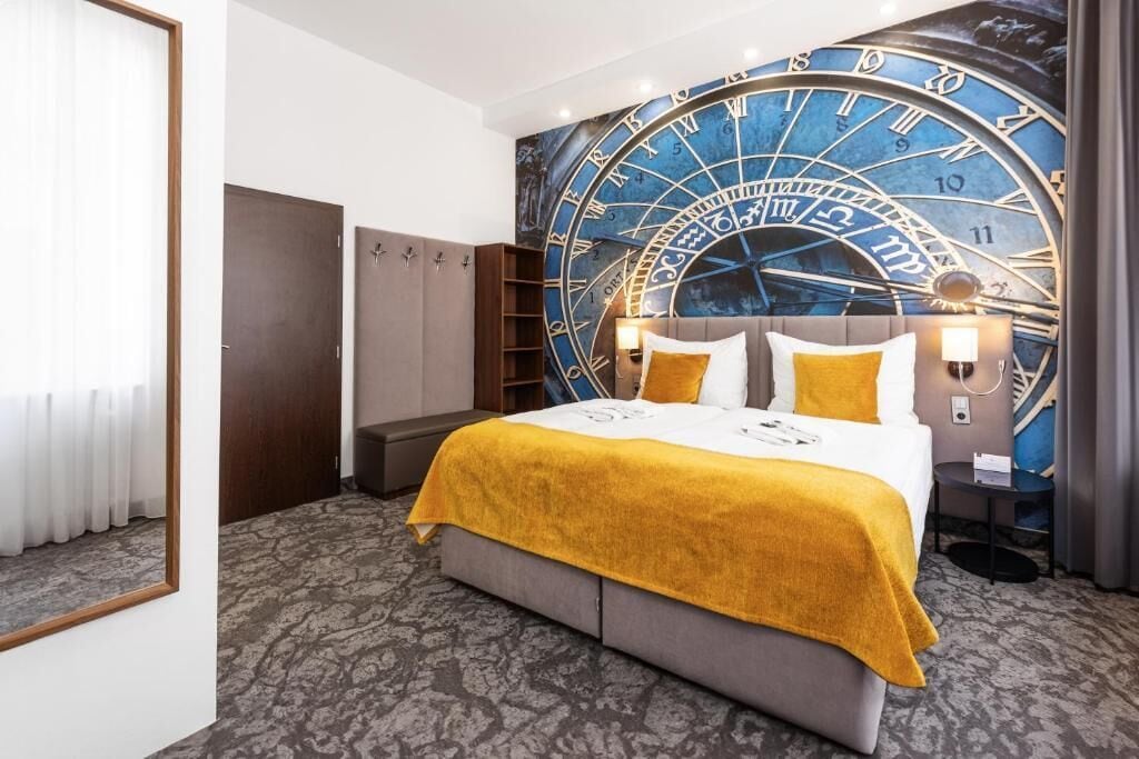 Ein Hotelzimmer mit einer großen Wanduhr, einem Bett mit gelber Decke, einem Kleiderschrank und einem Nachttisch mit einer Lampe.