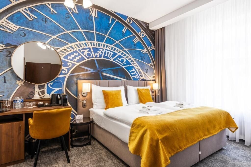 Une chambre d’hôtel avec une grande horloge murale, un lit avec une literie blanche et un plaid jaune, un bureau avec une chaise et un miroir.