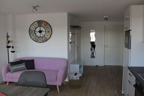 A modern living space with a sofa, a round wall clock, a small table, and a kitchen area. - Penthouse - Moments of Happiness on Rügen (Putbus)