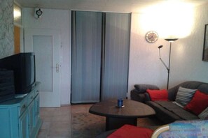 Een woonkamer met een televisie op een kast, een bank met rode kussens, een ronde salontafel en een vloerlamp.