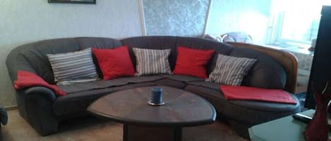 A living room with a dark-colored sectional sofa, red and striped cushions, a round coffee table with a blue object on it, and a green side table with a vase.