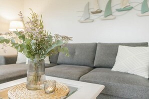A living room with a grey sofa, a white cushion, a glass vase with greenery, and a small glass on a woven placemat. - Holiday apartment in Nebel with swimming pool (Nebel)