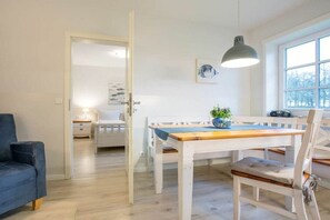 Ein Zimmer mit einem Holztisch, einem blauen Sofa, einem weißen Stuhl, einem Nachttisch mit Lampe und einem Fenster mit Blick auf Bäume.