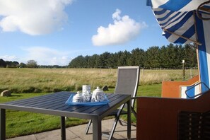 Eine Terrasse mit Gartenmöbeln und einem Tisch, gedeckt mit Teekannen und Tassen, mit Blick auf ein Wiesenfeld und Bäume.