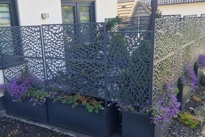 Ein schwarzer Metallzaun mit aufwendigen Ausschnitten, begleitet von schwarzen Pflanzkästen mit violetten Blumen.