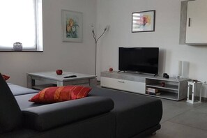 Uma sala de estar moderna com um sofá, uma televisão de tela plana, um armário branco e um abajur.