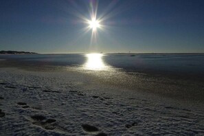Ein Strand mit Fußspuren, die zum Wasser führen, ein klarer Himmel und eine tief stehende Sonne.