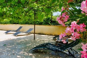 Een zonnig terras met lounge fauteuils en een tafel, omgeven door weelderig groen en bloeiende planten.
