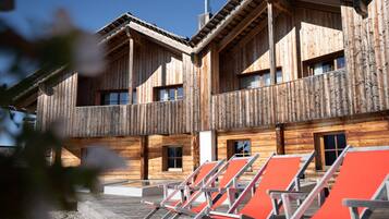 A wooden building with multiple balconies and windows, featuring outdoor seating areas with orange chairs.