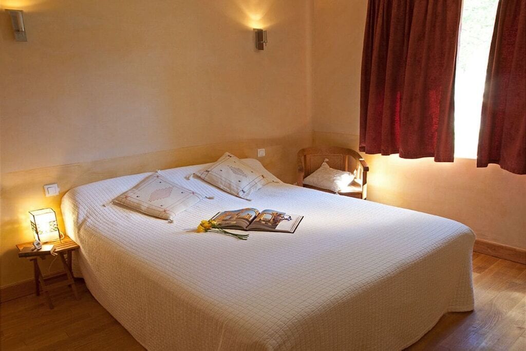 Cama bem arrumada com travesseiros e um livro, mesa de cabeceira com abajur e janela com cortinas.