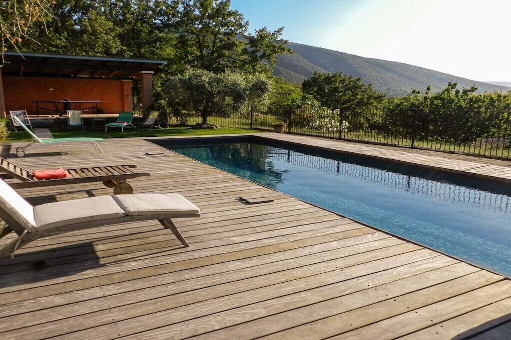 A wooden deck with a swimming pool, lounge chairs, and a view of a mountain.