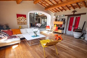Ein Wohnzimmer mit Sofa, Couchtisch und einem orangefarbenen Sessel. An der Wand hängt ein Gemälde und eine Tür ist mit roten Vorhängen drapiert.