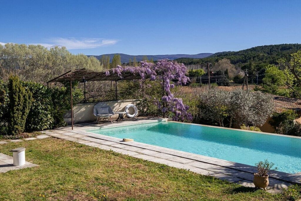 A swimming pool with a pergola covered in purple flowers, a barbecue area, and a view of hills.
