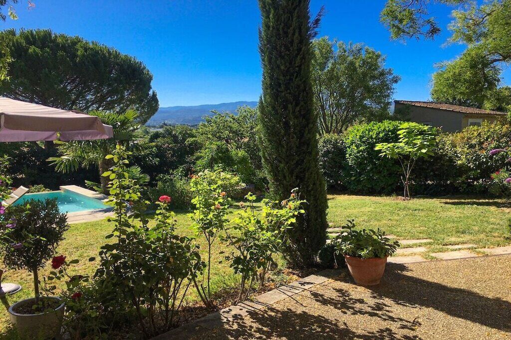 Un jardín con piscina, un árbol alto y un edificio al fondo.