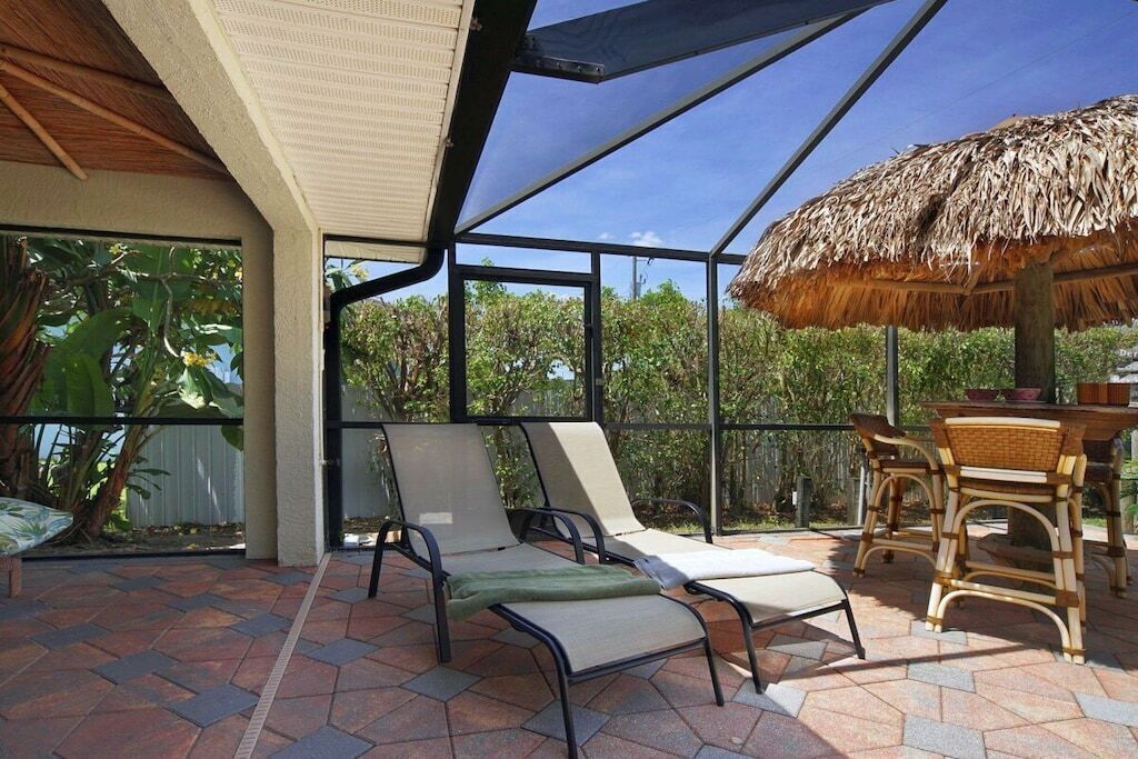 A covered outdoor seating area with lounge chairs and a table, surrounded by glass walls and a thatched roof.