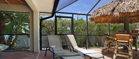 Ein überdachter Außenbereich mit Lounge-Möbeln und einem Tisch, umgeben von Glaswänden und einem Reetdach.